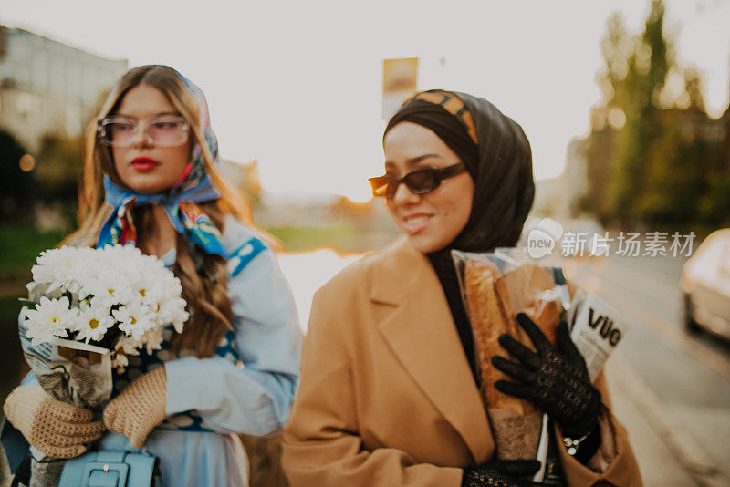 A modern European woman and a Muslim woman with a hijab walking the streets of the city dressed in clothes from the 19th century while carrying newspapers, flowers and bread in their hands.